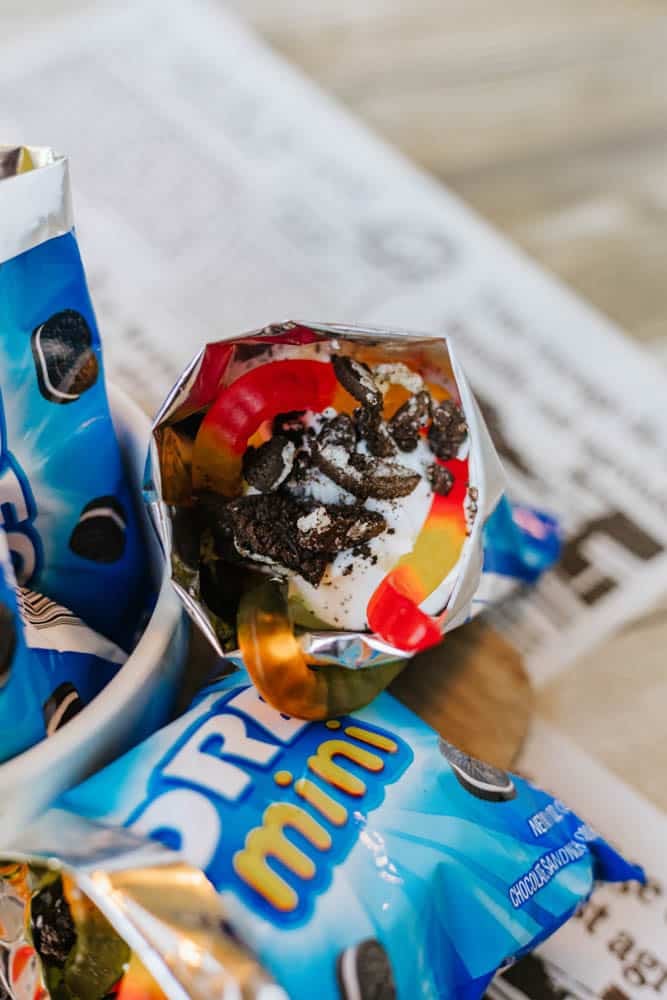 An opened mini Oreo cookie snack bag filled with whipped cream, crushed Oreos, and gummy worms, sitting on a newspaper-covered table.