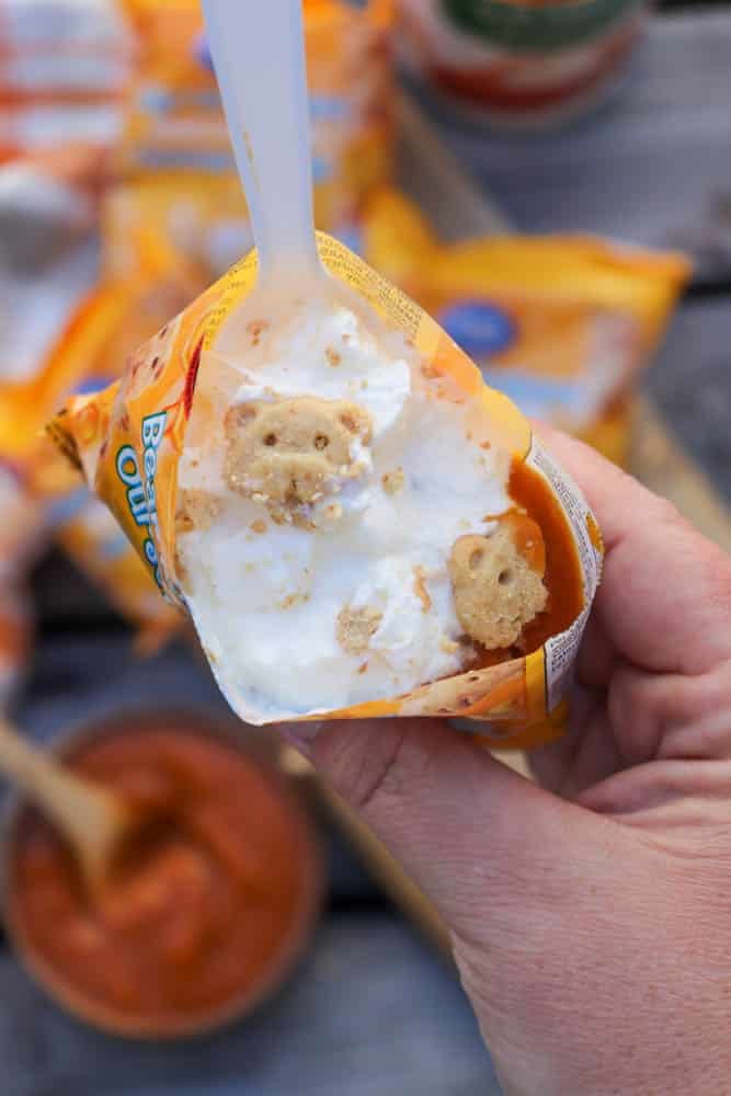 A hand holds an open snack-size bag of bear-shaped graham cookies topped with whipped cream and pumpkin filling, with a white plastic spoon inside. A bowl of pumpkin filling is seen in the background.