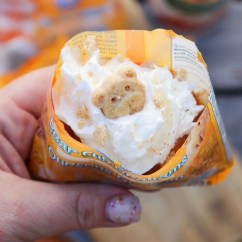 A hand holds an open packet of bear-shaped graham cookies filled with whipped cream, with one cookie visible on top. A blurred wooden surface and snacks are in the background.