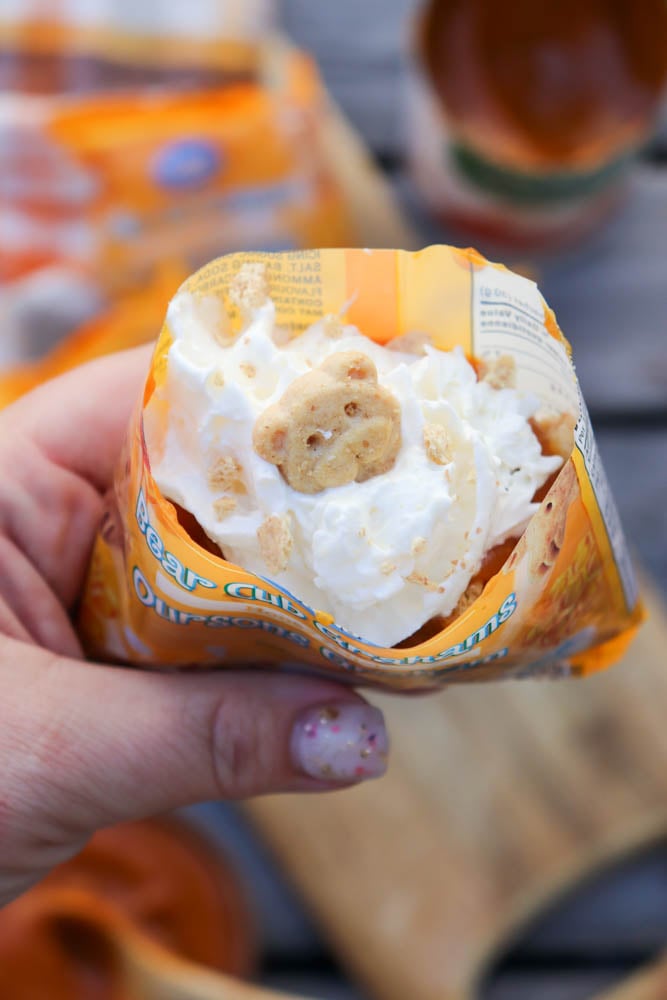 A hand holds an open packet of bear-shaped graham cookies filled with whipped cream, with one cookie visible on top. A blurred wooden surface and snacks are in the background.