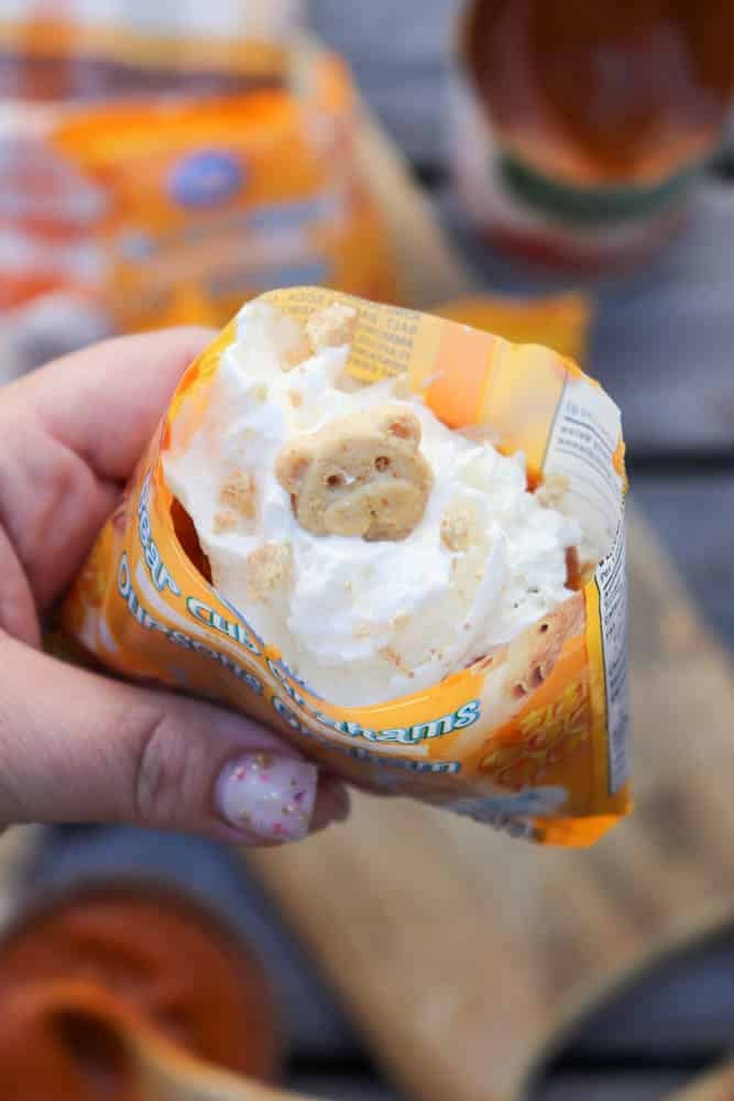 A hand holds an opened bag of Teddy Grahams cookies filled with whipped cream, topped with a teddy bear-shaped cookie. There is a wooden surface and blurred food items in the background.