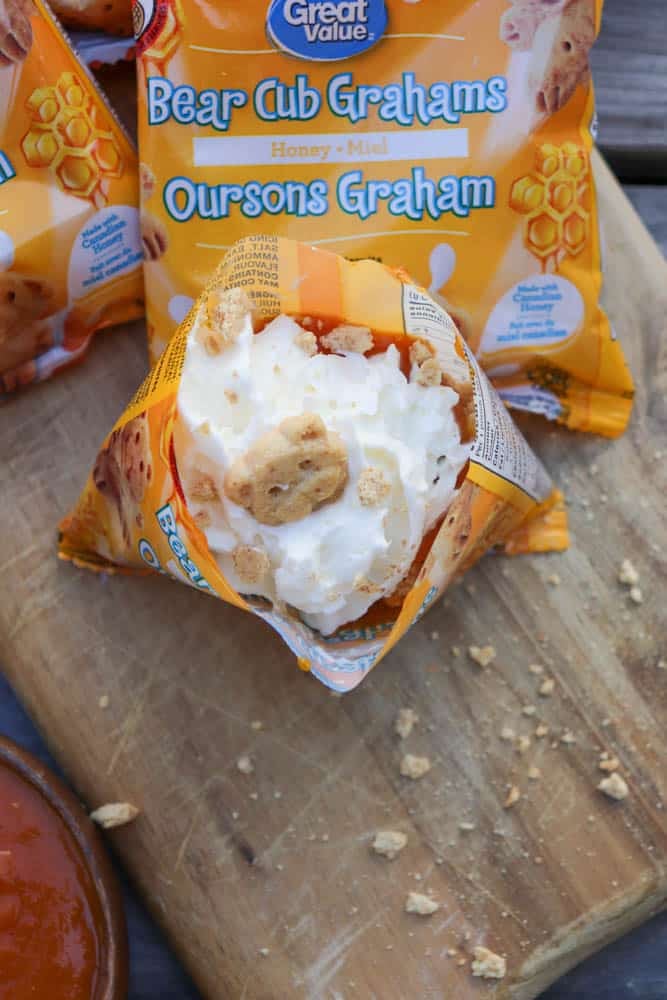 An open bag of Bear Cub Grahams filled with whipped cream and topped with cookie pieces sits on a wooden surface, with another unopened bag and some cookie crumbs nearby.