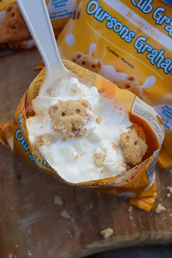 A plastic spoon scoops vanilla ice cream from an opened bag of honey graham bear-shaped cookies, with cookie pieces mixed into the ice cream. The yellow packaging is partially visible.