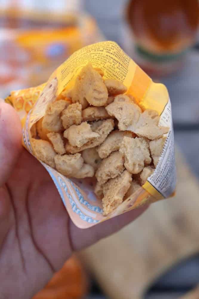 A hand holds an open bag filled with small, broken pieces of crunchy, beige-colored snacks or cookies. The bag has a yellow interior and printed text. The background is blurred.
