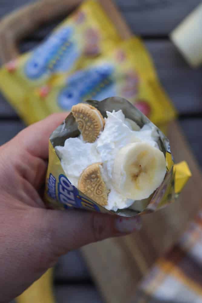 A hand holds an opened mini Oreo cookie bag filled with whipped cream, banana slices, and golden mini Oreo cookies, with cookie packages blurred in the background.