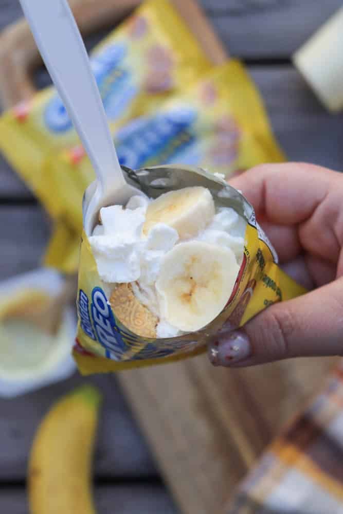 A hand holds an open mini Oreo cookie packet filled with whipped cream and banana slices, using a white plastic spoon. A banana, cutting board, and more Oreos are blurred in the background.
