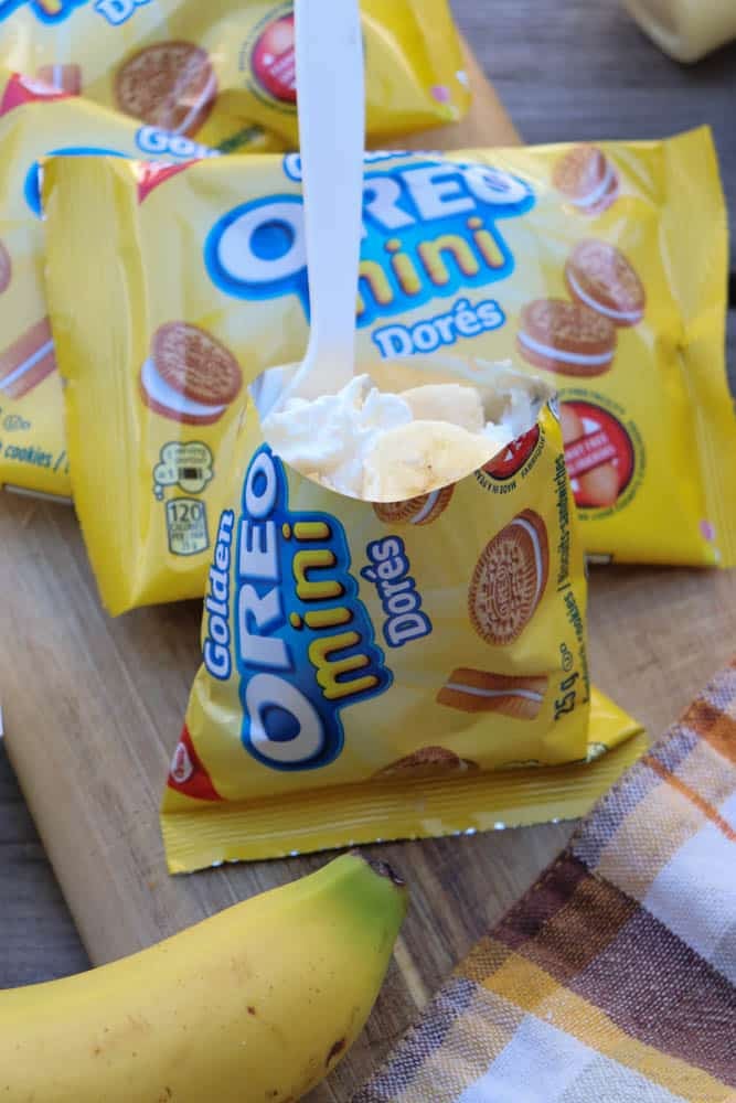 A small yellow bag of Golden Oreo Mini cookies is opened and filled with whipped cream and banana slices, with a white plastic spoon inside. The bag sits on a wooden surface next to a banana and a plaid cloth.