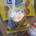 A packet of Golden Oreo Mini cookies is open with whipped cream and banana slices inside. A white plastic spoon is inserted. Unopened cookie packs, a banana, and a plaid napkin are nearby on a wooden surface.