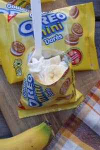 A packet of Golden Oreo Mini cookies is open with whipped cream and banana slices inside. A white plastic spoon is inserted. Unopened cookie packs, a banana, and a plaid napkin are nearby on a wooden surface.