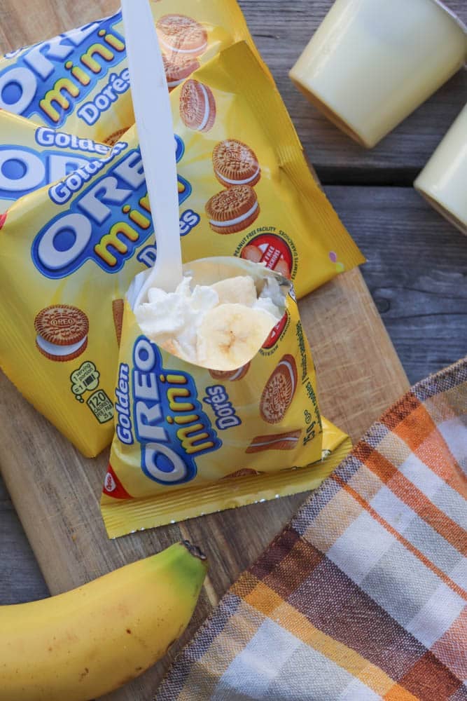A package of Golden Oreo Mini cookies is opened and filled with whipped cream and banana slices, placed on a wooden board next to a banana, a plaid napkin, and pudding cups.