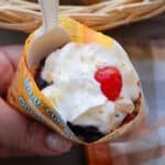 A hand holds a small, open snack package filled with whipped cream, a cherry, and crumbled topping, served with a white plastic spoon. There is a checkered napkin and a wicker basket in the background.