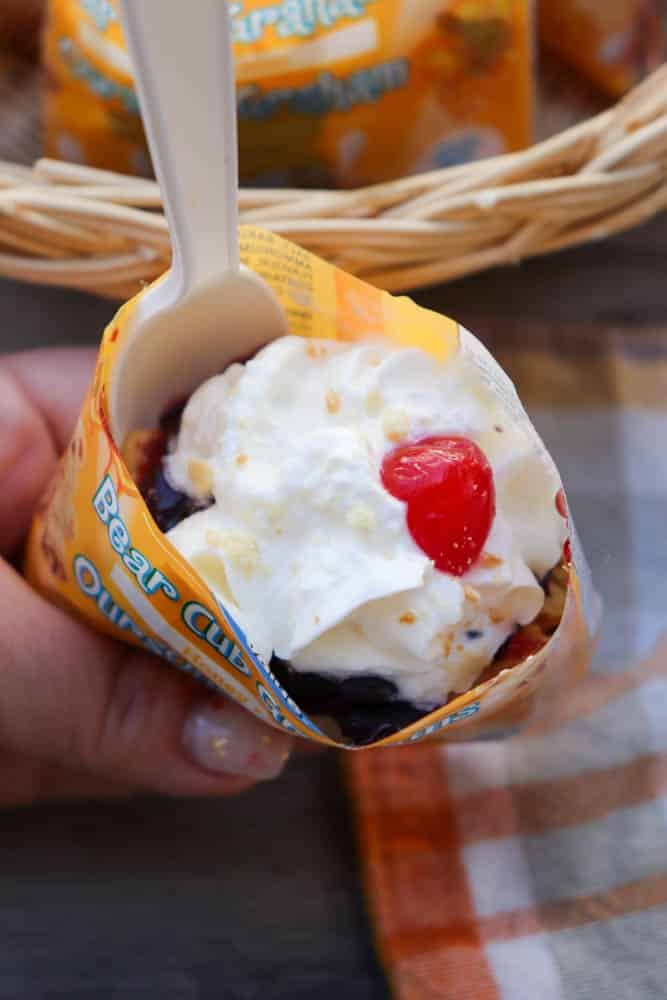 A hand holds a small, open snack package filled with whipped cream, a cherry, and crumbled topping, served with a white plastic spoon. There is a checkered napkin and a wicker basket in the background.