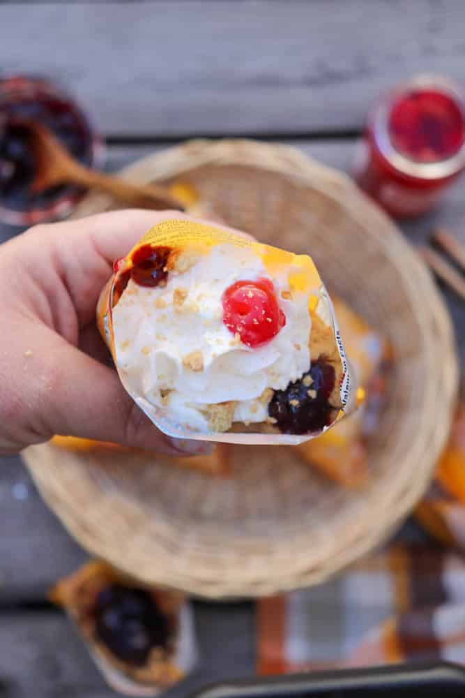 A hand holds a waffle cone filled with whipped cream, cherry, and blueberry toppings, with a sprinkle of crumbs. There’s a woven basket and jars of toppings on a wooden table in the blurred background.