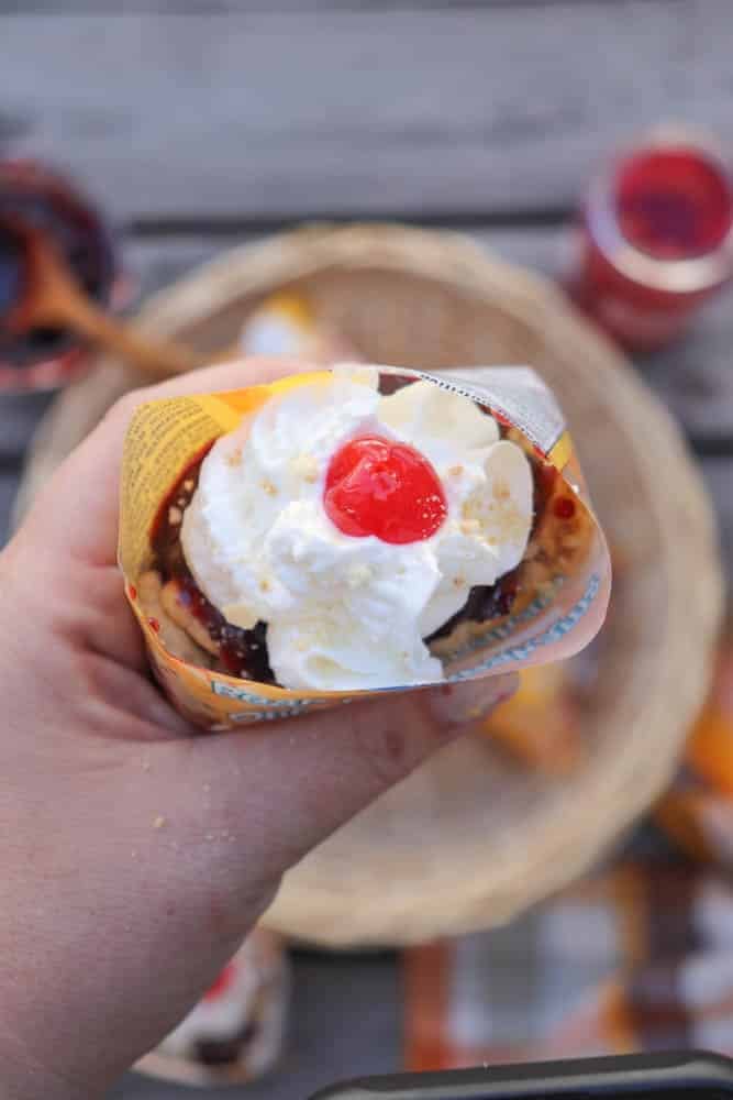 A hand holds an ice cream cone topped with whipped cream and a cherry. The cone is partially wrapped in a colorful wrapper, and a basket with more cones is blurred in the background.