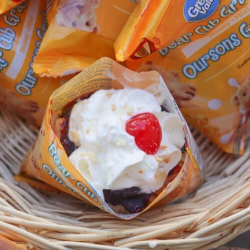 A waffle cone filled with ice cream, whipped cream, and a cherry, surrounded by Great Value Bear Club Grahams snack packs in a wicker basket.