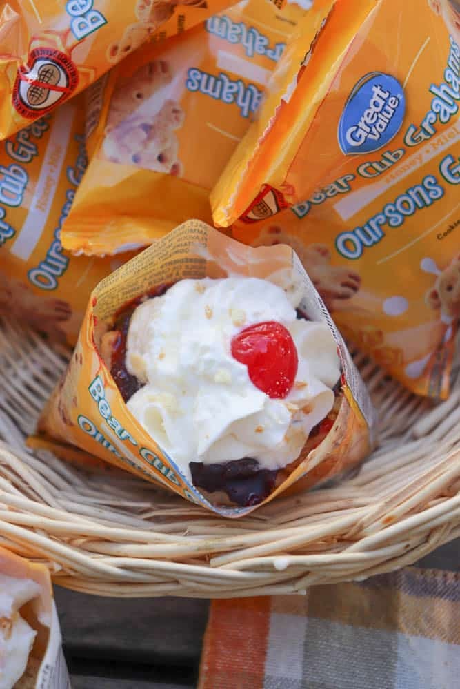 A waffle cone filled with ice cream, whipped cream, and a cherry, surrounded by Great Value Bear Club Grahams snack packs in a wicker basket.