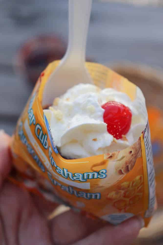 A close-up of a hand holding an opened bag of graham snacks filled with whipped cream and topped with a maraschino cherry, with a white plastic spoon inserted.