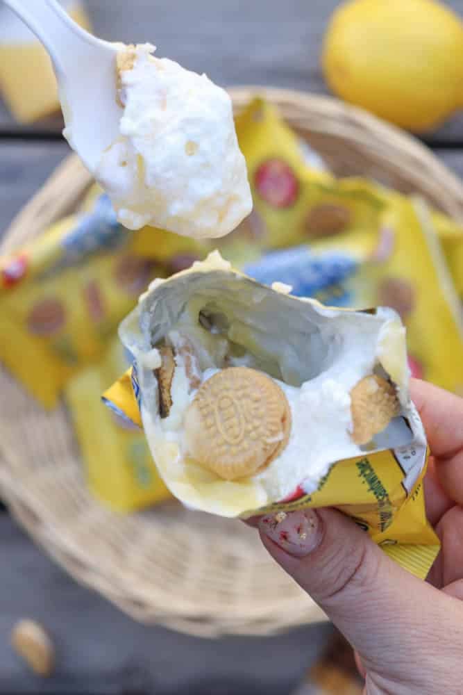 A hand holds an open snack-sized bag of Golden Oreo cookies filled with creamy dessert, while a spoon scoops some out. A basket with more cookie bags and a lemon are in the background.