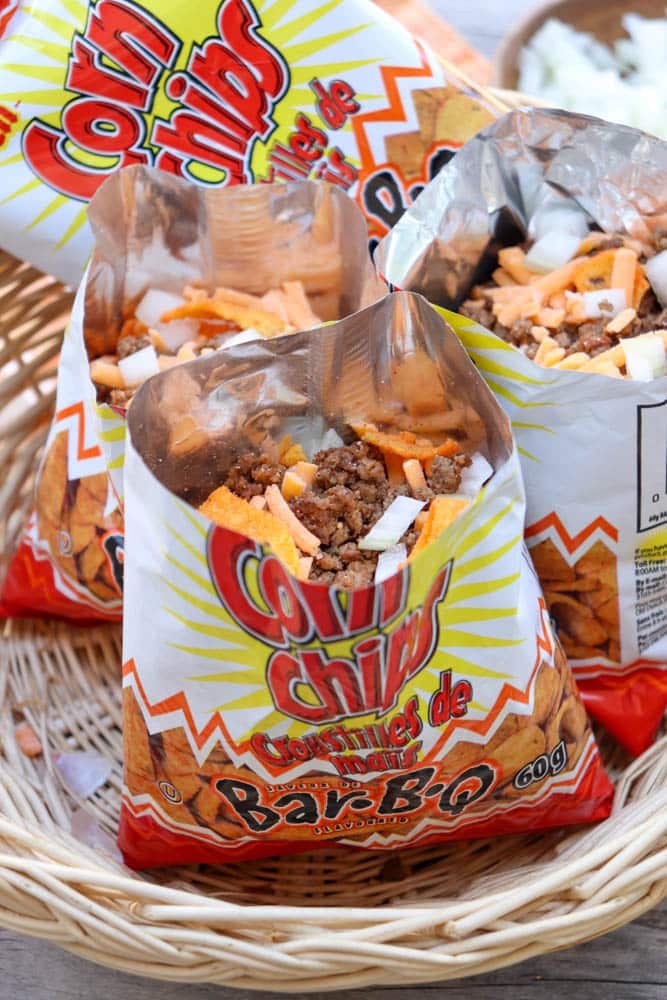 Open bags of corn chips filled with ground beef, shredded cheese, and chopped onions are arranged in a wicker basket, creating a portable snack or meal.