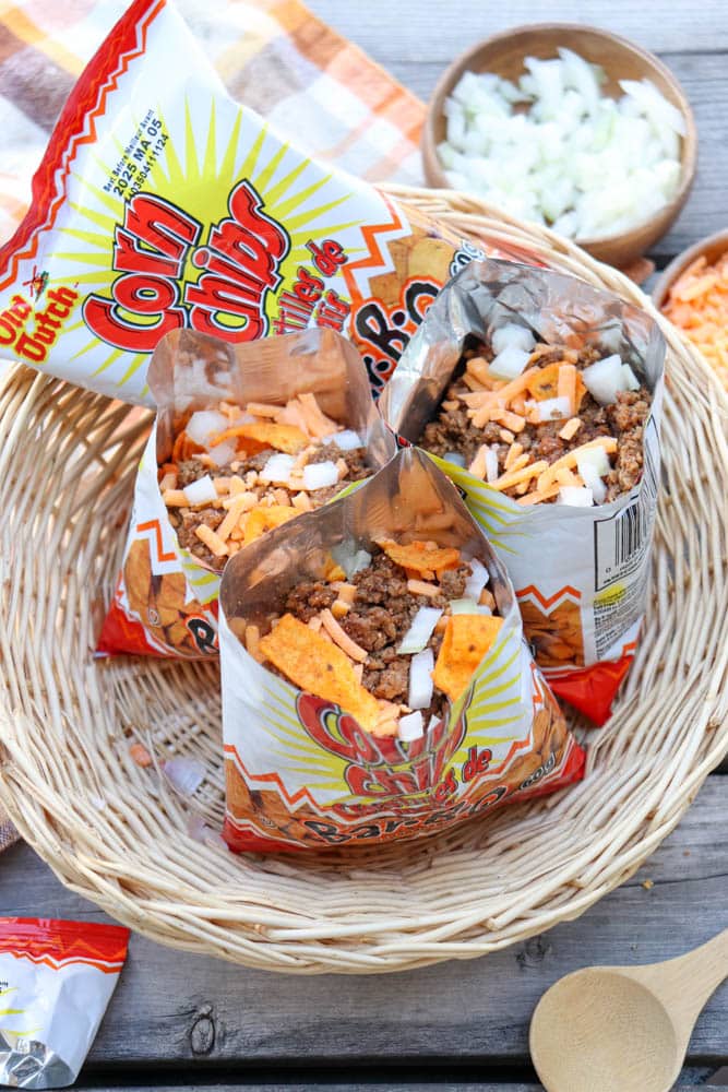 Three opened snack-sized bags of Corn Chips filled with seasoned ground beef, shredded cheese, and chopped onions sit in a wicker basket. Bowls of diced onions and cheese are nearby on a wooden surface.