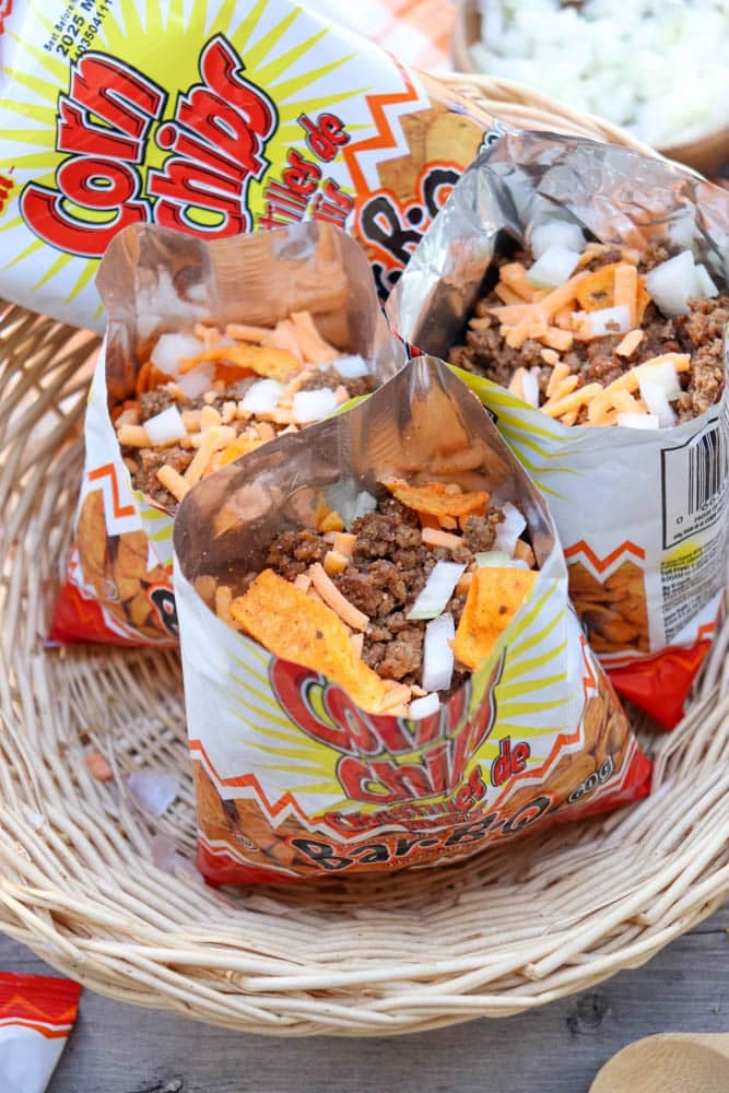 Three opened bags of corn chips filled with ground beef, shredded cheese, and chopped onions sit in a woven basket, with a partially empty corn chips bag in the background.
