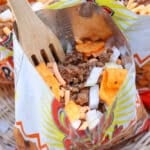A close-up of a small opened chip bag filled with ground beef, chopped onions, shredded cheese, and orange chips, with a wooden fork inside. The bag rests in a wicker basket.
