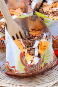 A close-up of a small opened chip bag filled with ground beef, chopped onions, shredded cheese, and orange chips, with a wooden fork inside. The bag rests in a wicker basket.