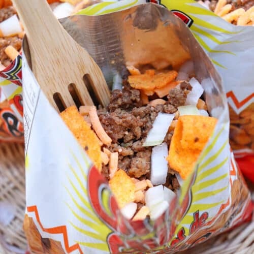 A close-up of a small opened chip bag filled with ground beef, chopped onions, shredded cheese, and orange chips, with a wooden fork inside. The bag rests in a wicker basket.