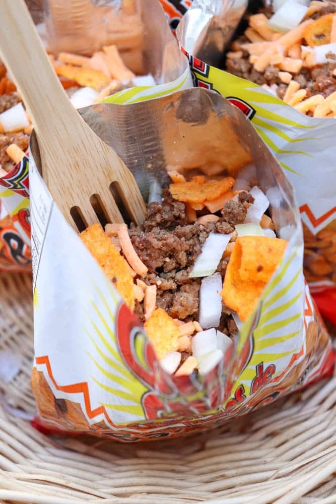 A close-up of a small opened chip bag filled with ground beef, chopped onions, shredded cheese, and orange chips, with a wooden fork inside. The bag rests in a wicker basket.