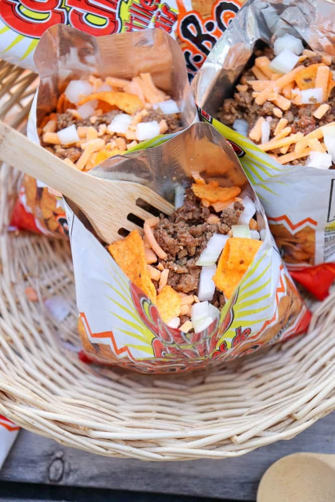 An open bag of chips filled with ground beef, shredded cheese, chopped onions, and a wooden fork, displayed in a woven basket with other similar chip bags in the background.