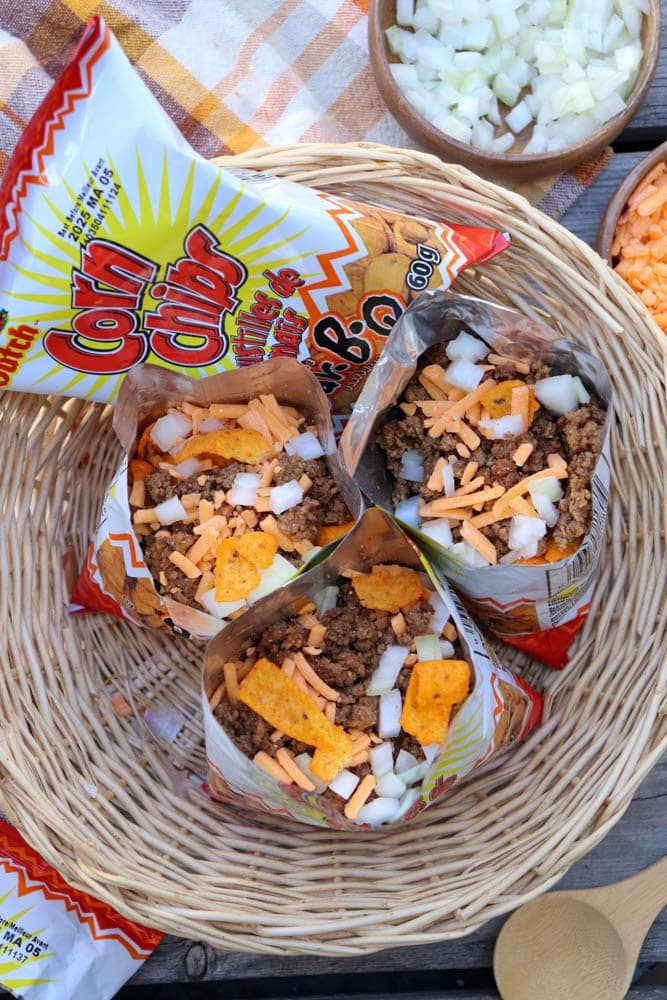 Three open bags of corn chips in a woven basket, each filled with taco meat, shredded cheese, and chopped onions. Bowls of diced onions and shredded cheese are nearby on a checkered cloth.