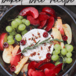 A cast iron skillet filled with a wheel of brie cheese, sliced plums, apples, green grapes, cherries, and pecans, garnished with a rosemary sprig, sits over a campfire.