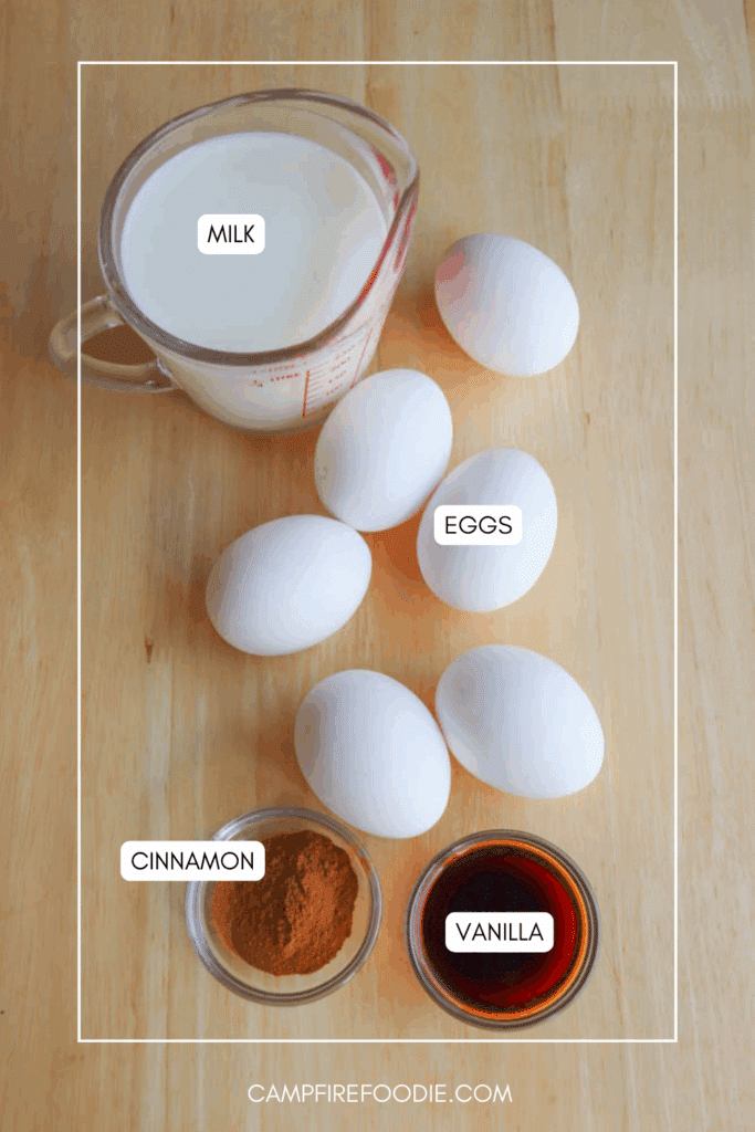 A measuring cup of milk, five white eggs, a small bowl of ground cinnamon, and a small bowl of vanilla extract arranged on a wooden surface. Labels indicate each ingredient.