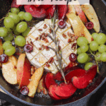 A cast iron skillet with baked brie topped with nuts and herbs, surrounded by sliced apples, plums, green grapes, and cherries. Text overlay reads Camping Skillet Brie Recipe and campfirefoodie.com.