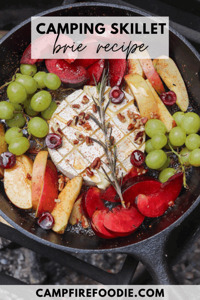 A cast iron skillet with baked brie topped with nuts and herbs, surrounded by sliced apples, plums, green grapes, and cherries. Text overlay reads Camping Skillet Brie Recipe and campfirefoodie.com.