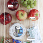 A flat lay of ingredients on a wooden surface, including green grapes, plums, red apples, a wheel of brie cheese, a box of fresh rosemary, and a bag of pecans. Labels identify each item.