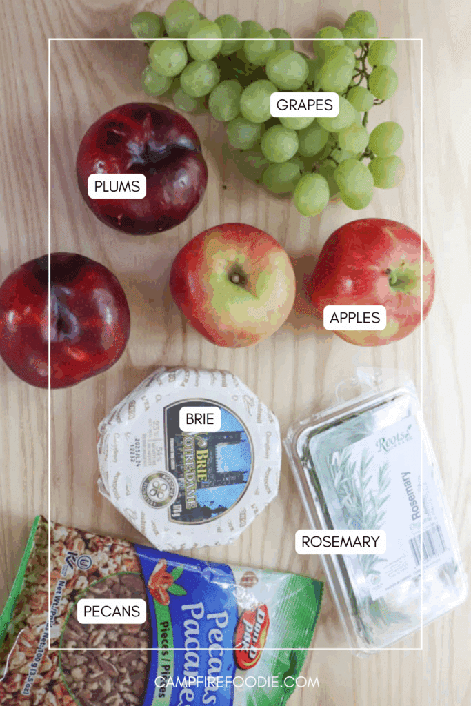 A flat lay of ingredients on a wooden surface, including green grapes, plums, red apples, a wheel of brie cheese, a box of fresh rosemary, and a bag of pecans. Labels identify each item.