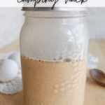 A mason jar filled with a french toast mixture sits on a wooden table, with two white eggs and a wooden spoon in the background. Text reads “FRENCH TOAST camping hack” and “CAMPFIREFOODIE.COM.”.