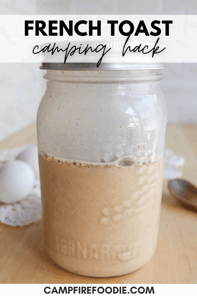 A mason jar filled with a french toast mixture sits on a wooden table, with two white eggs and a wooden spoon in the background. Text reads “FRENCH TOAST camping hack” and “CAMPFIREFOODIE.COM.”.