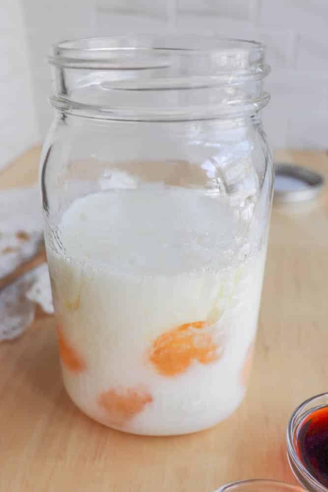 A glass jar filled with milk and several cracked eggs submerged inside, sitting on a wooden surface with a cloth and a small bowl of dark liquid nearby.