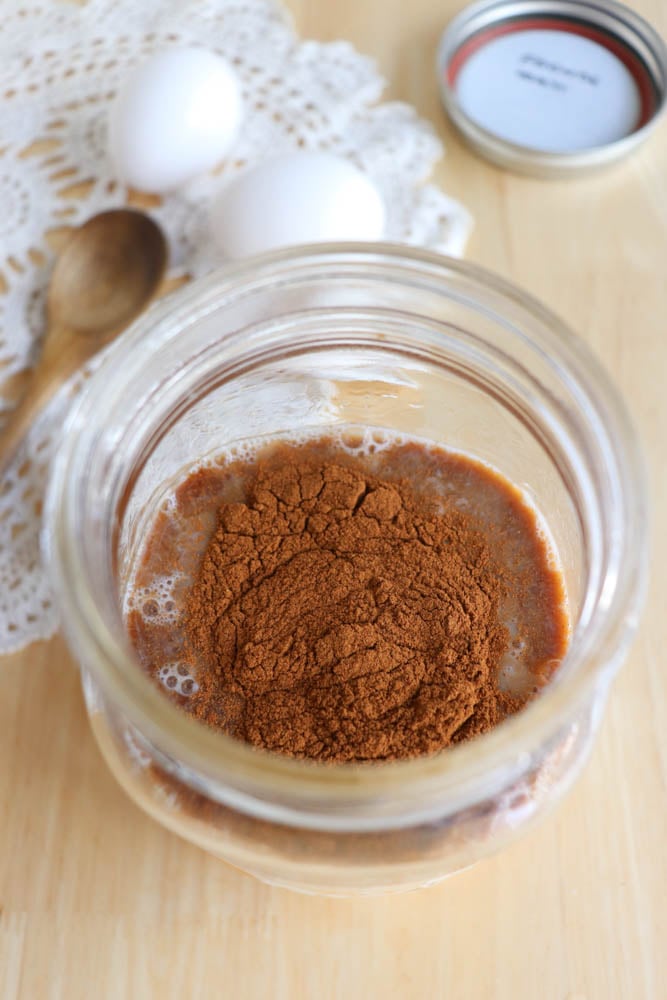 A glass jar filled with a light brown liquid topped with a mound of cinnamon powder. Nearby are two white eggs, a wooden spoon, a mason jar lid, and a lace doily on a wooden surface.