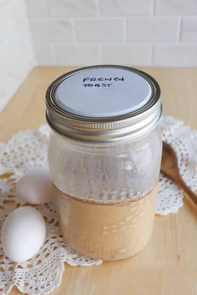 A glass jar labeled French Toast sits on a crocheted doily with two eggs and a wooden spoon nearby, on a light wooden surface against a white tiled background.