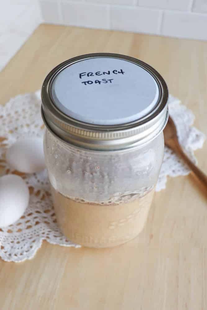 A glass mason jar labeled French Toast sits on a wooden table with a white doily, two eggs, and a wooden spoon nearby. The jar appears to contain a liquid mixture.
