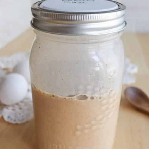 A mason jar with a metal lid containing a beige liquid sits on a wooden table next to two white eggs, a doily, and a wooden spoon, with a white tile wall in the background.