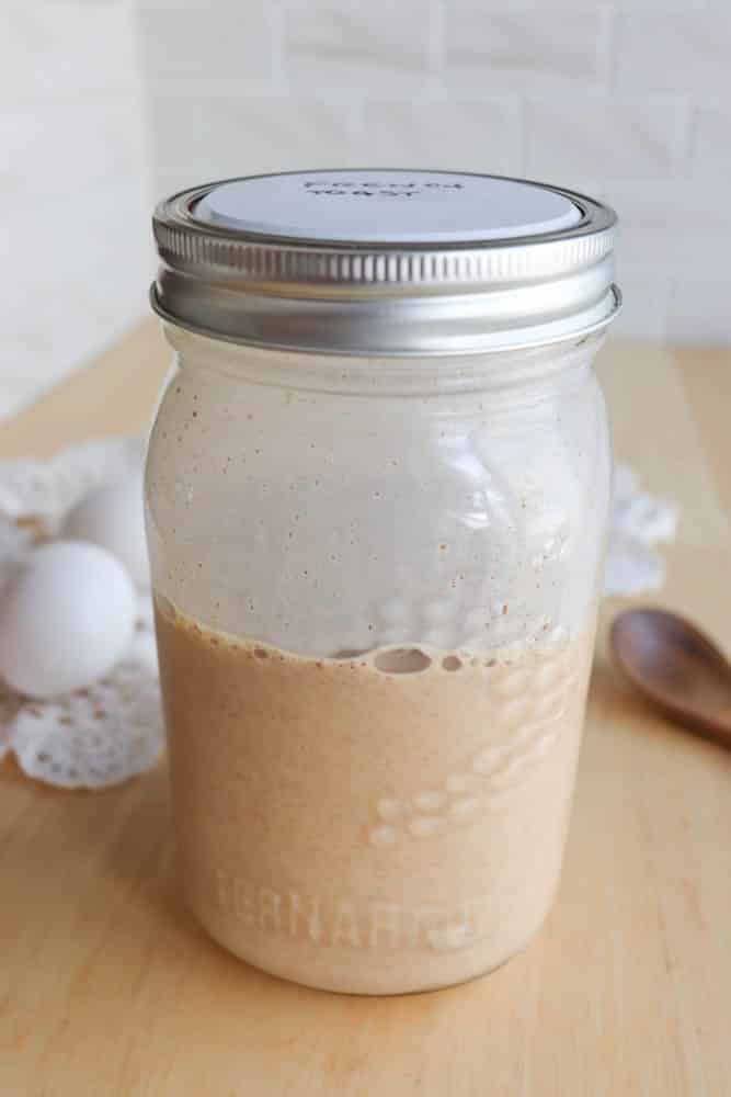 A mason jar with a metal lid containing a beige liquid sits on a wooden table next to two white eggs, a doily, and a wooden spoon, with a white tile wall in the background.