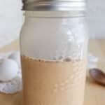 A mason jar filled with a creamy, light brown liquid sits on a wooden table. Two eggs, a lace doily, and a wooden spoon are in the background.