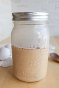 A mason jar filled with a creamy, light brown liquid sits on a wooden table. Two eggs, a lace doily, and a wooden spoon are in the background.