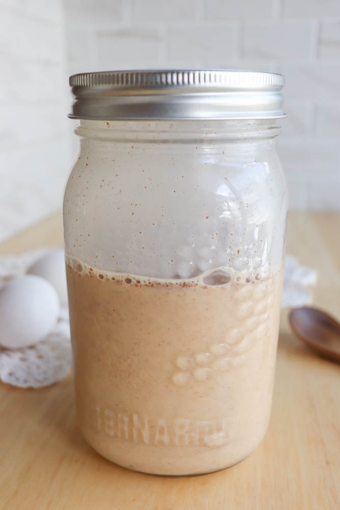 A mason jar filled with a creamy, light brown liquid sits on a wooden table. Two eggs, a lace doily, and a wooden spoon are in the background.