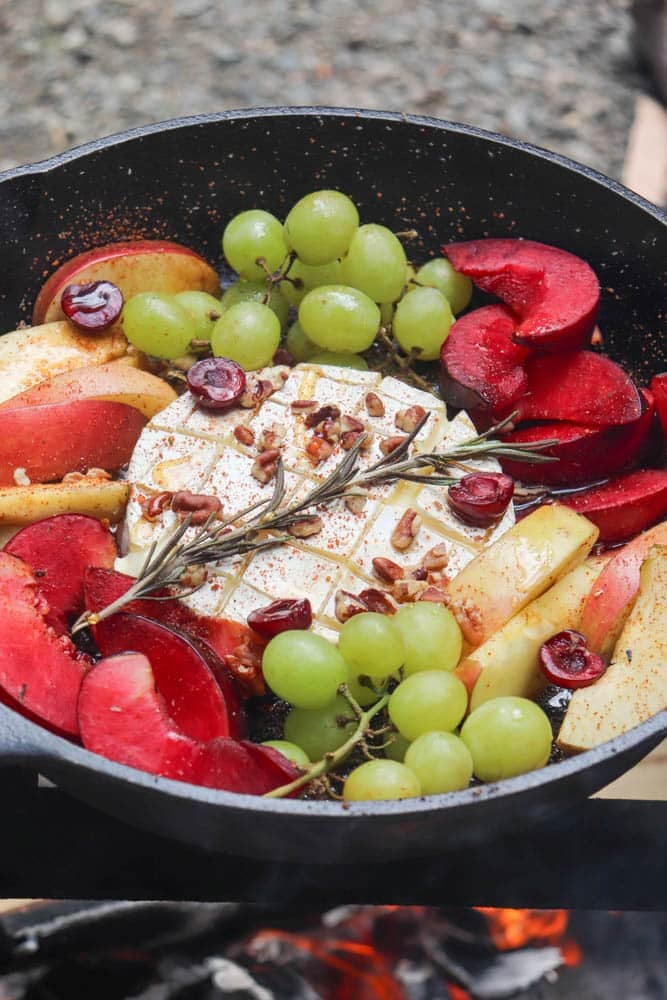 A cast iron skillet filled with a round of brie cheese, surrounded by sliced apples, plums, green grapes, cherries, chopped nuts, and rosemary, all atop an outdoor grill with embers visible below.