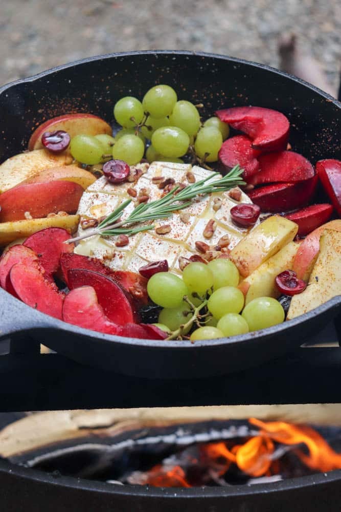 A cast iron skillet filled with baked brie, green grapes, apple and plum slices, fresh rosemary, and chopped pecans sits over an open campfire.